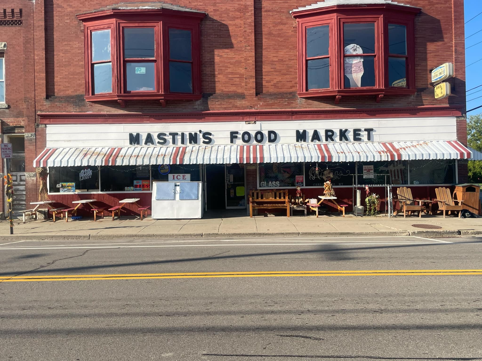 Mastin's Food Market storefront on Main Street in Canaseraga, NY — red-and-white striped awning over a brick facade, vintage MASTIN'S FOOD MARKET sign, picnic tables and Adirondack chairs out front